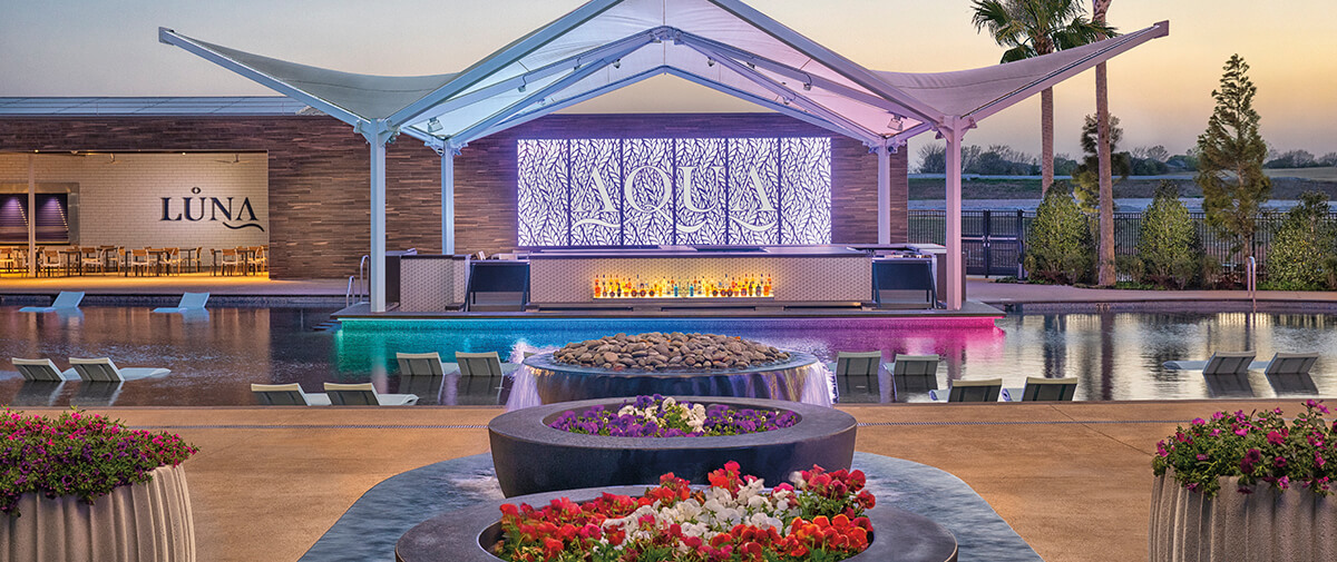 Poolside AQUA Bar with a sculptural canopy, illuminated backdrop, and water loungers, flower planters in the foreground.