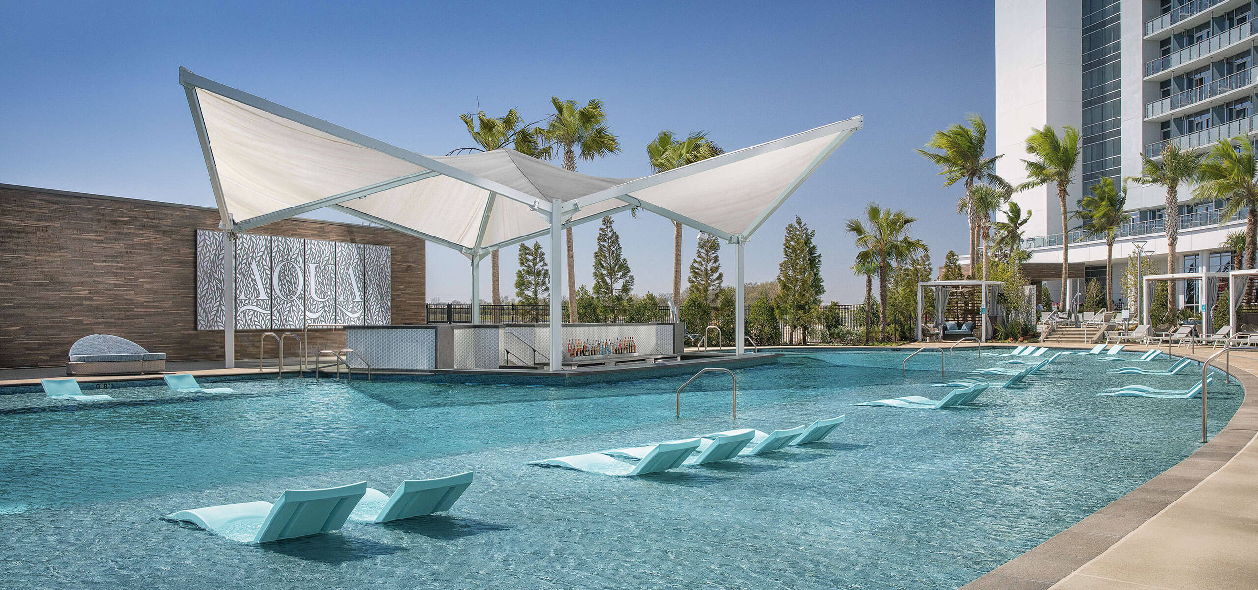 Expansive resort pool with white shade canopy, in-water loungers, palm trees, AQUA bar, and Choctaw hotel tower behind.