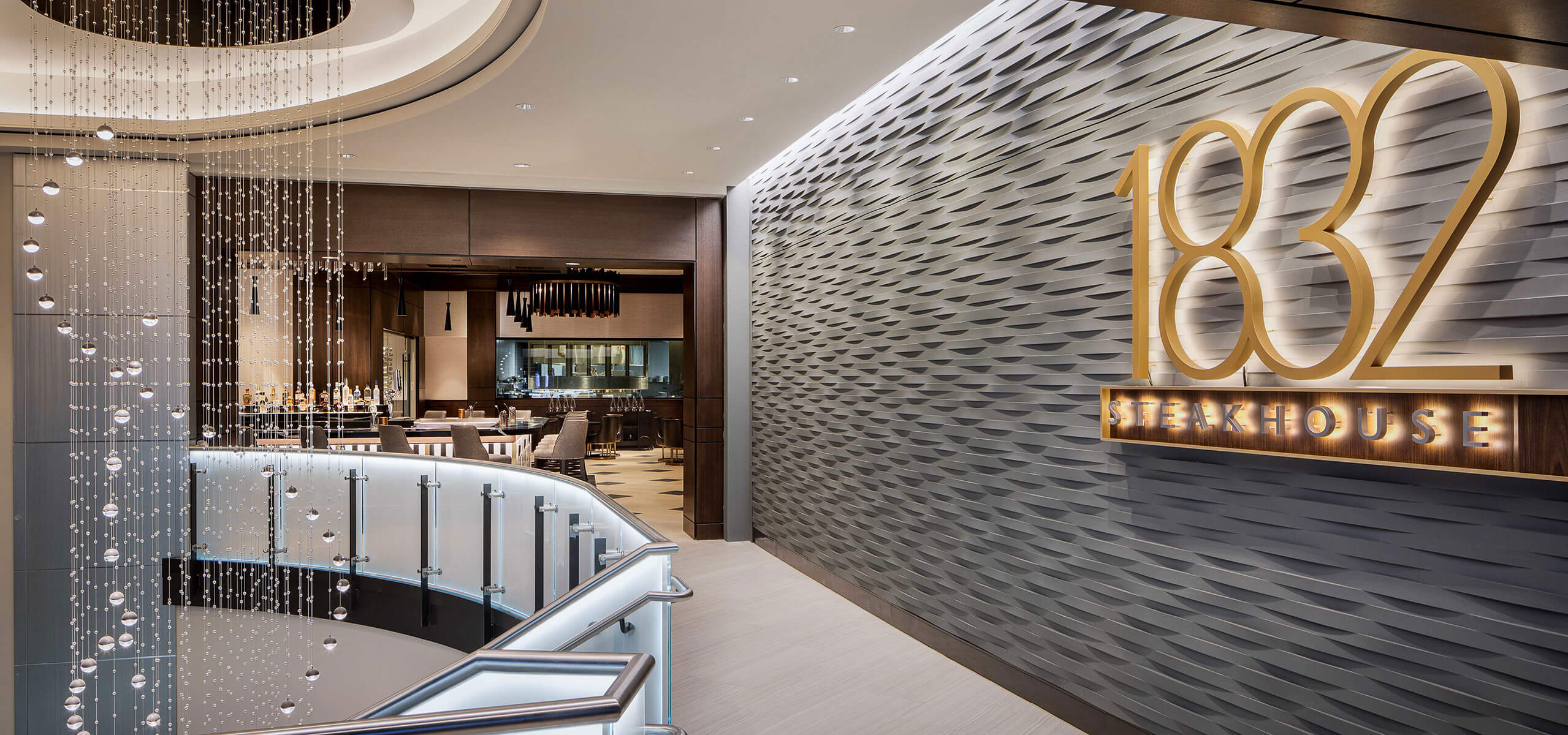 Entrance to 1832 Steakhouse with large illuminated sign, textured feature wall, glass-railed staircase, and crystal bead curtain.