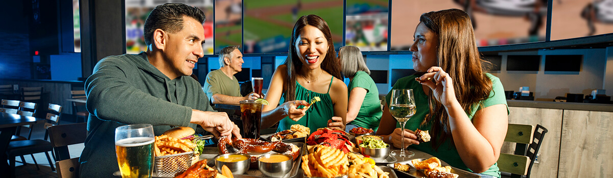 Three diners at Choctaw sports bar sharing burgers, ribs, fries, and drinks beneath a wall of big screens showing games.