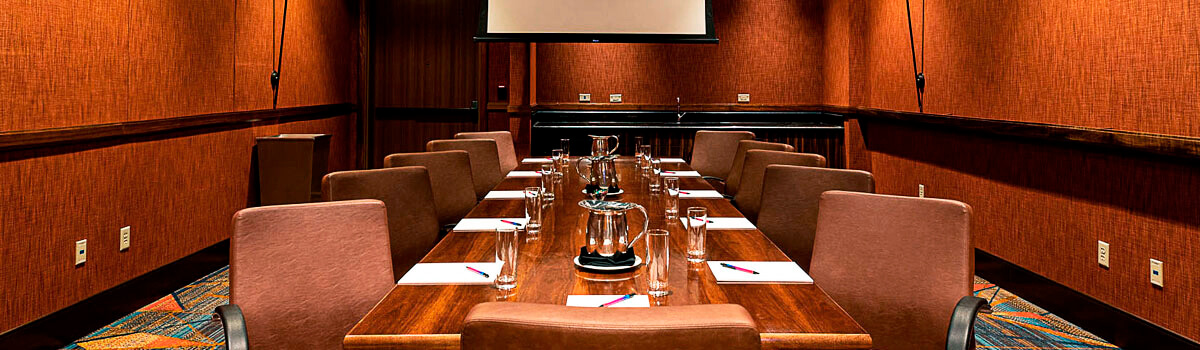 Cypress Boardroom with a long conference table, plush chairs, wood paneling, and a wall-mounted display under modern lighting.