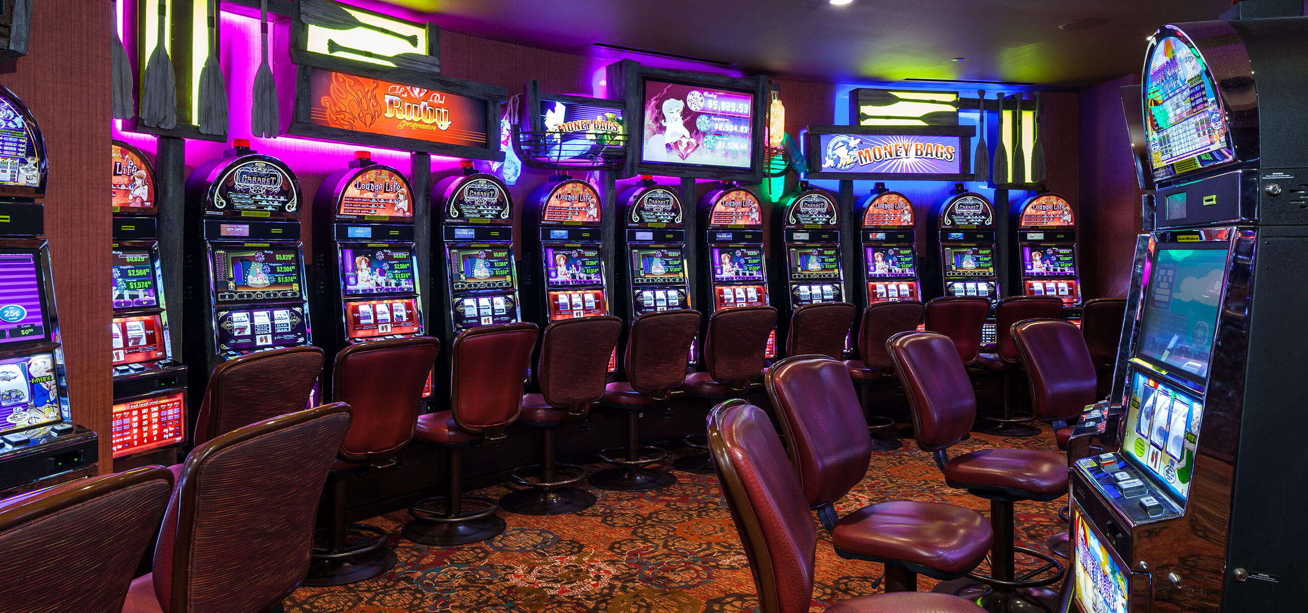 Row of classic slot machines with neon backlighting and red swivel chairs inside the casino